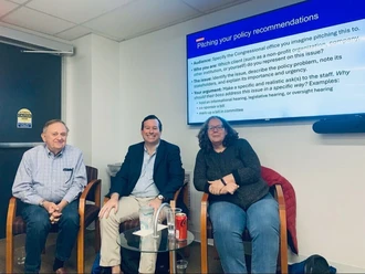 Three people sit in chairs in front of TV showing a presentation slide